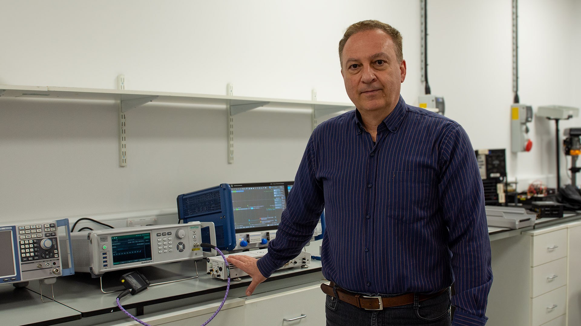 Individual standing in electronics lab beside testing equipment and monitors, with measurement devices, cables and wall shelving visible.