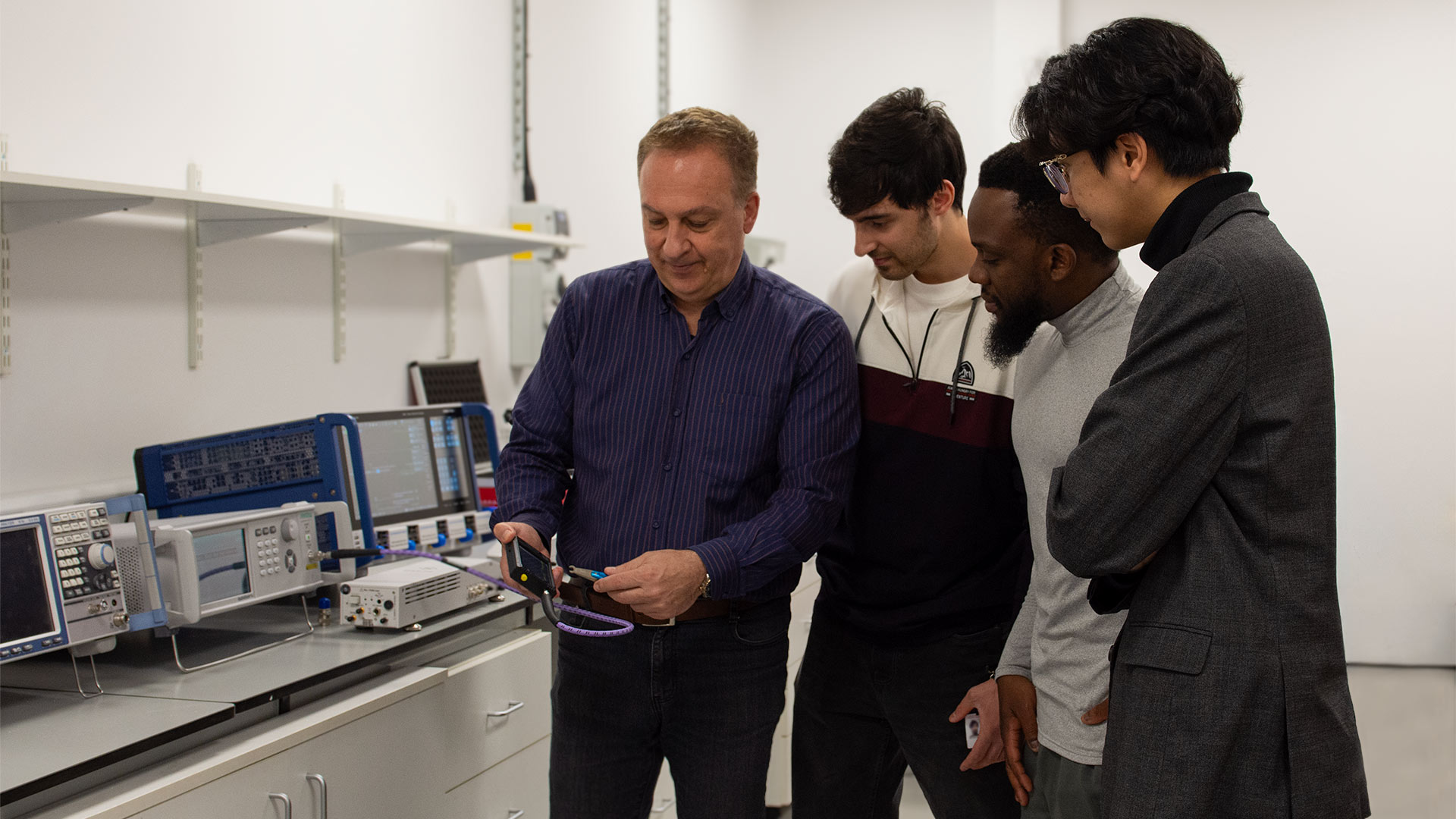 Group of four individuals in electronics lab gathered around testing equipment, as one demonstrates a small device connected by cable, with measurement instruments and monitors on workbench.
