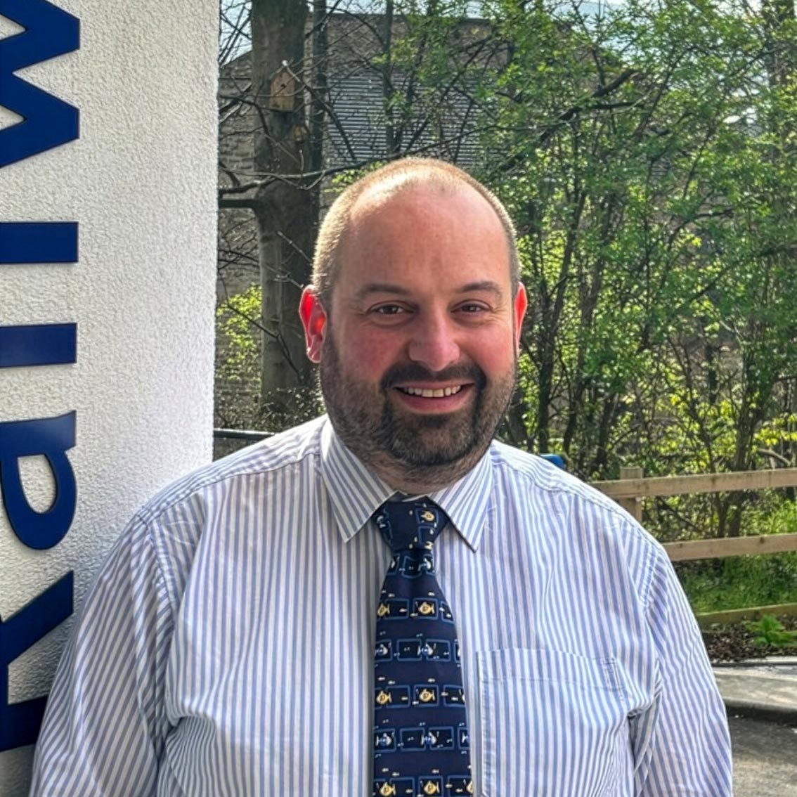 Individual in striped shirt and patterned tie stands outdoors near building signage, with trees and wooden fence in the background.
