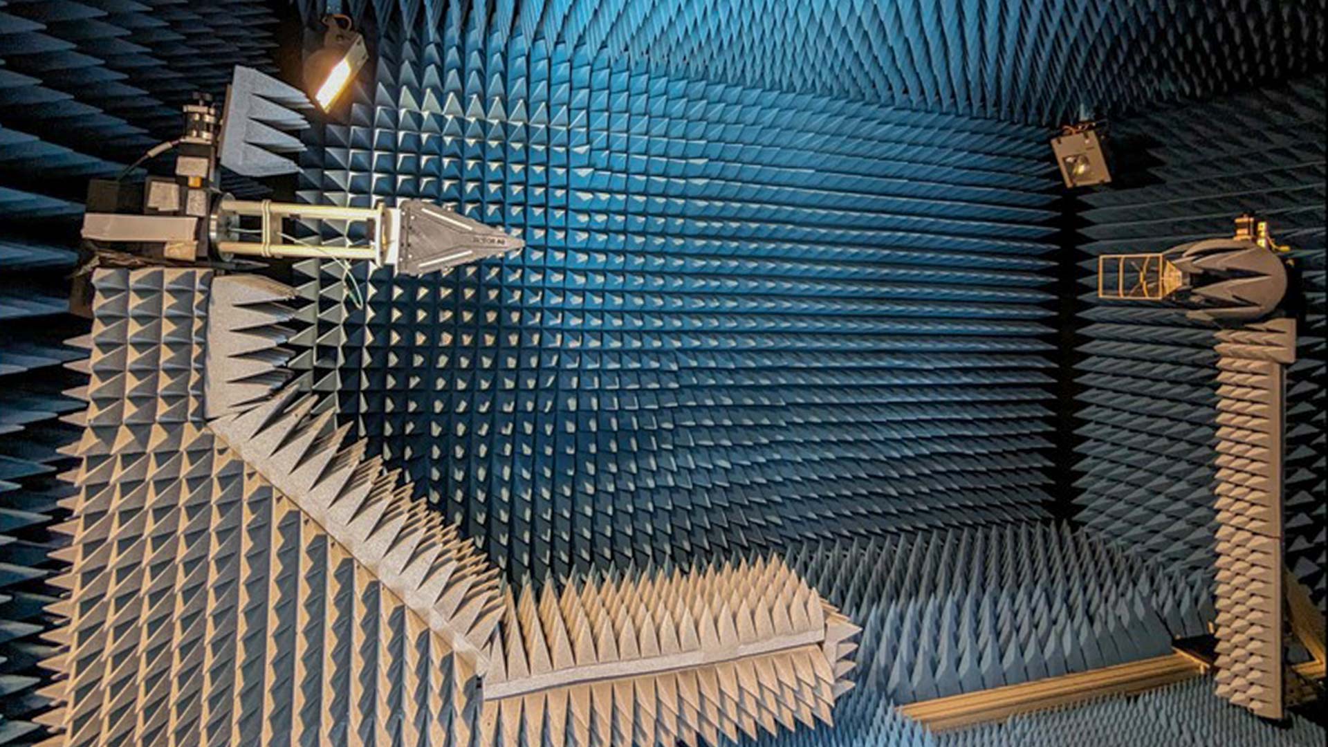 Anechoic chamber lined with blue acoustic foam wedges, containing mounted antennas and measurement equipment used for electromagnetic testing in a controlled environment.