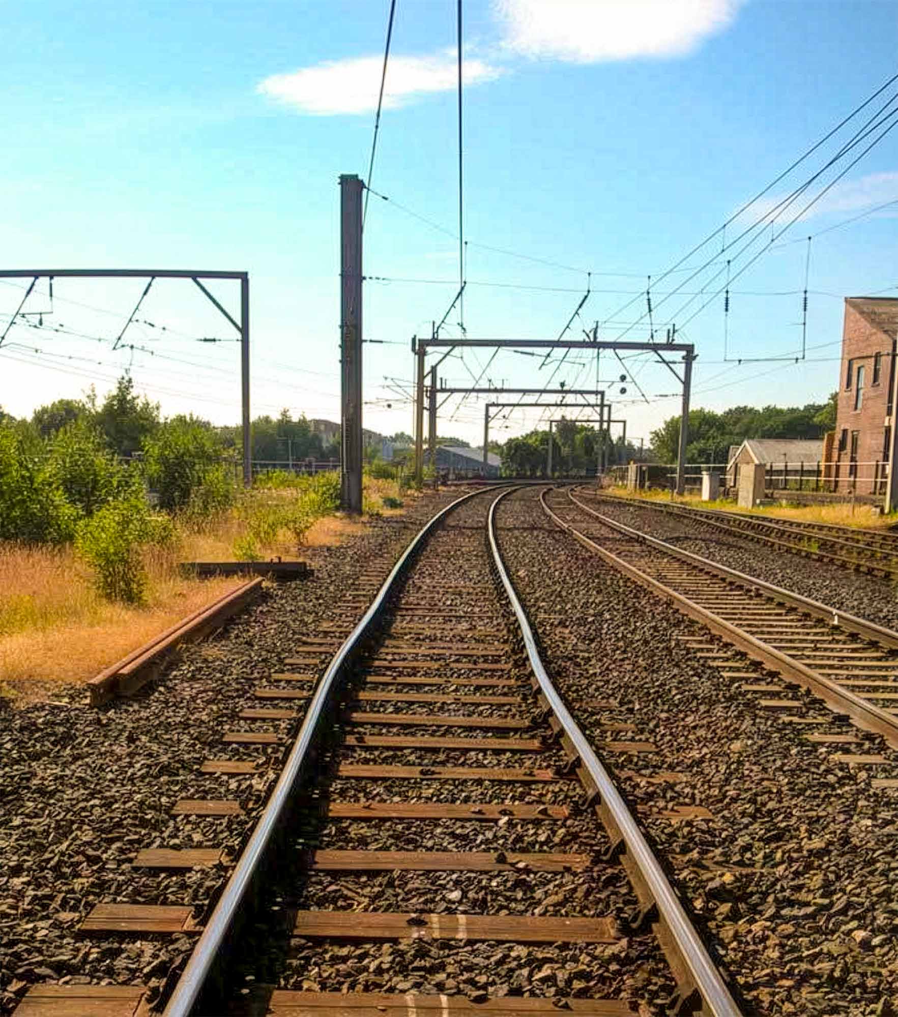 Railway tracks curve and buckle through outdoor landscape with overhead electrification lines, pylons and surrounding vegetation under blue sky.