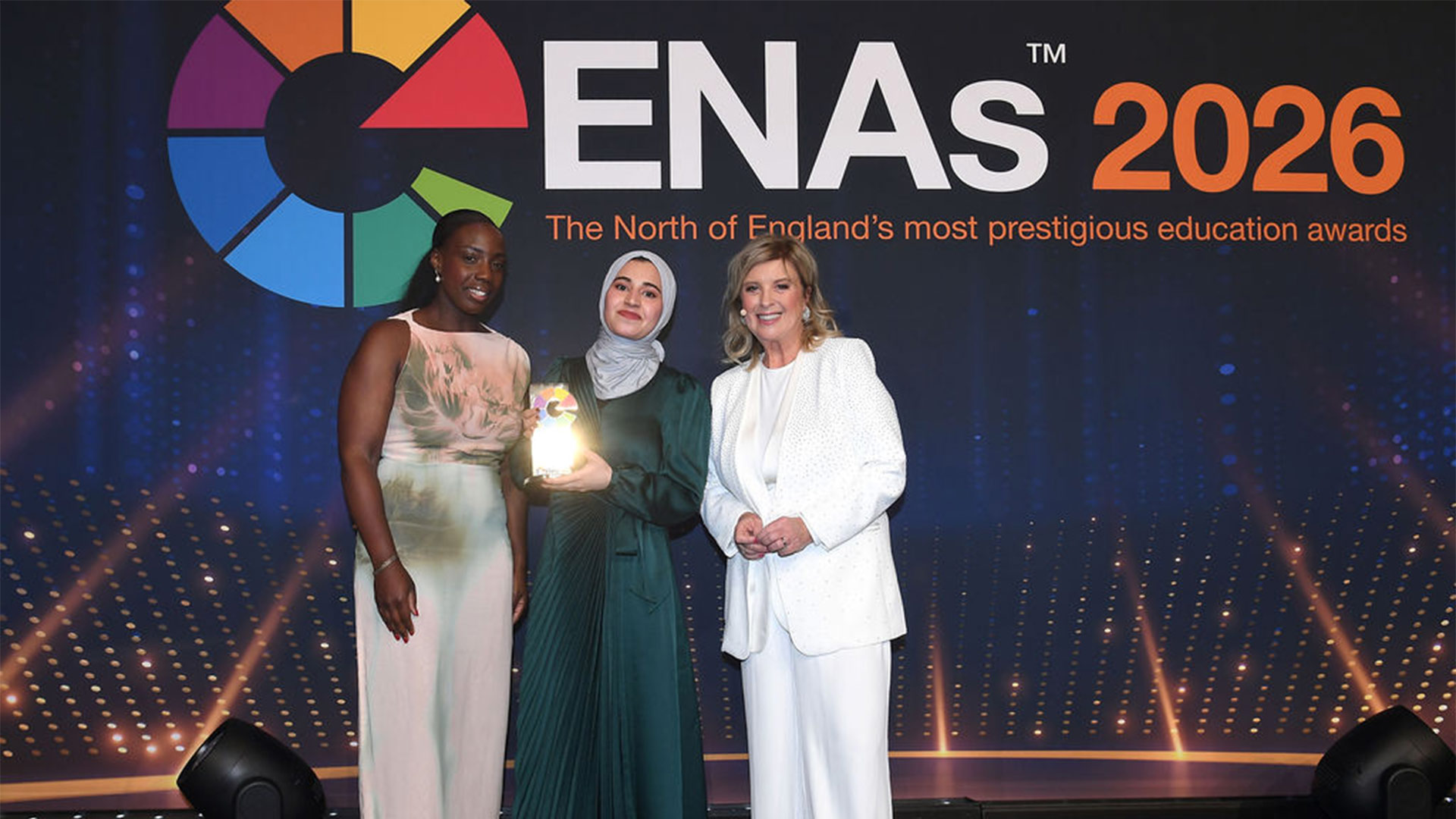 Three people in formalwear pose at the ENAs 2026, one holding a glass award trophy, standing on stage beneath prominent event logo and text.