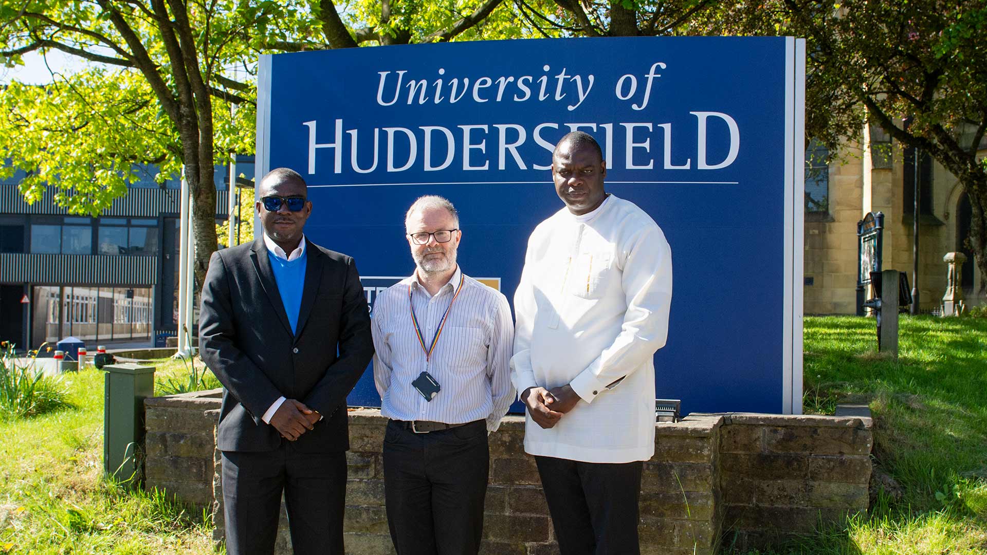 Three people stand outdoors in front of University of Huddersfield sign, with trees and campus buildings visible in bright sunlight.