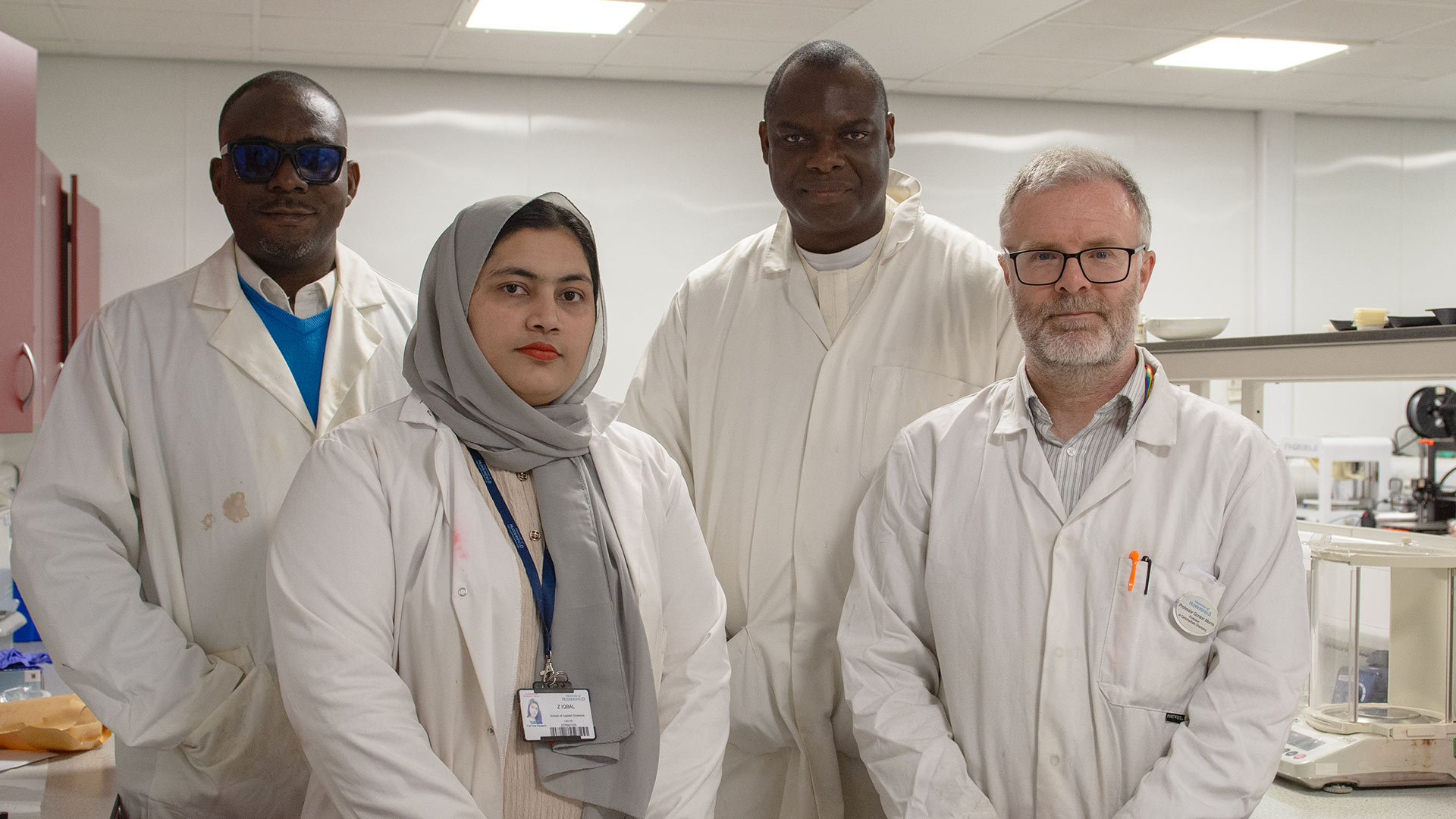Four researchers in laboratory coats stand together in a lab, with scientific equipment and workbenches visible in the background.