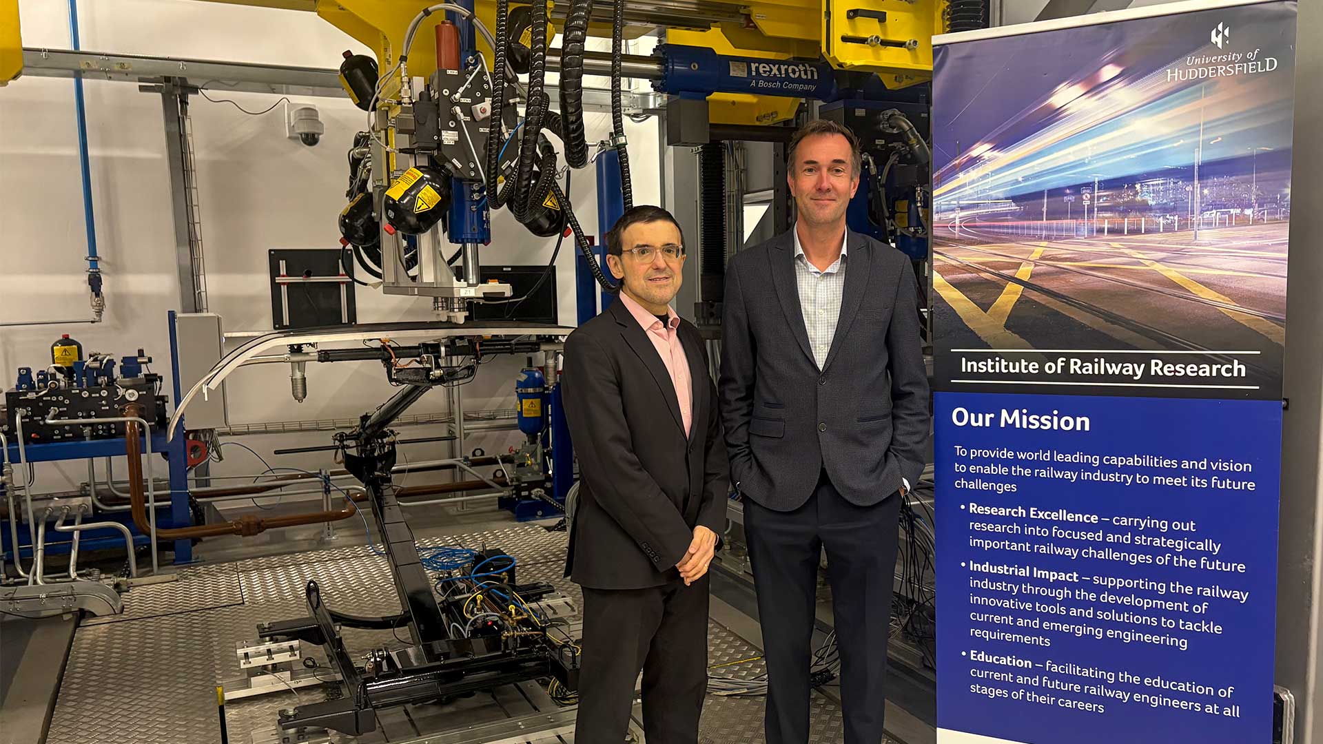 Two people in business attire stand inside engineering lab beside large testing machinery and University of Huddersfield Institute of Railway Research banner.