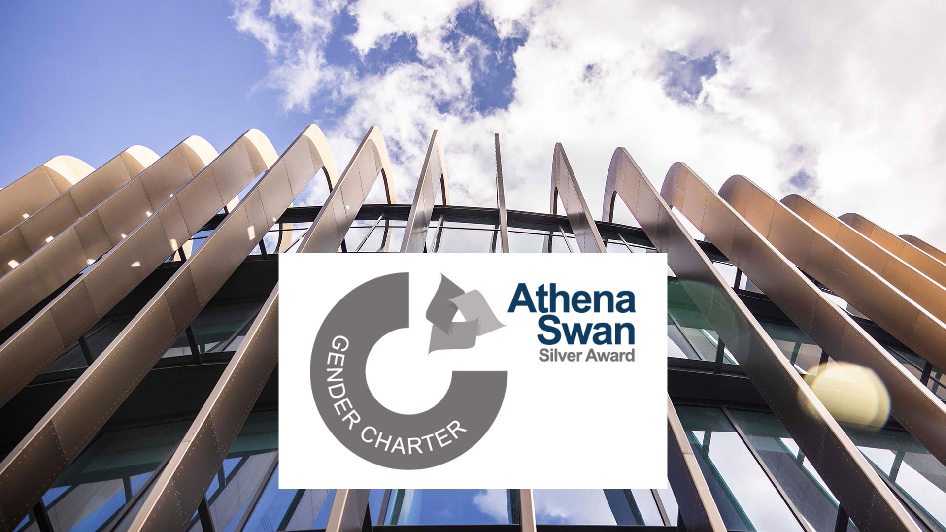 Modern university building with curved metal façade against blue sky, overlaid with Athena Swan Silver Award and Gender Charter logo, highlighting institutional commitment to gender equality.