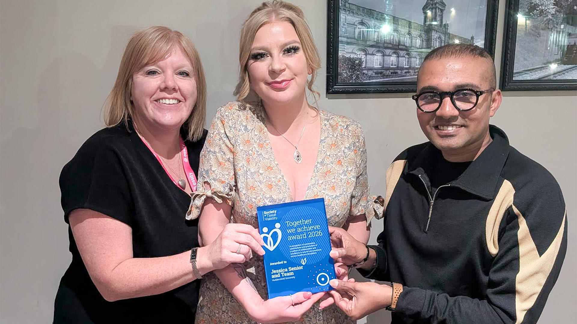 Three people posing indoors holding a blue “Together we achieve award 2026” certificate, with framed artwork visible on the wall behind.
