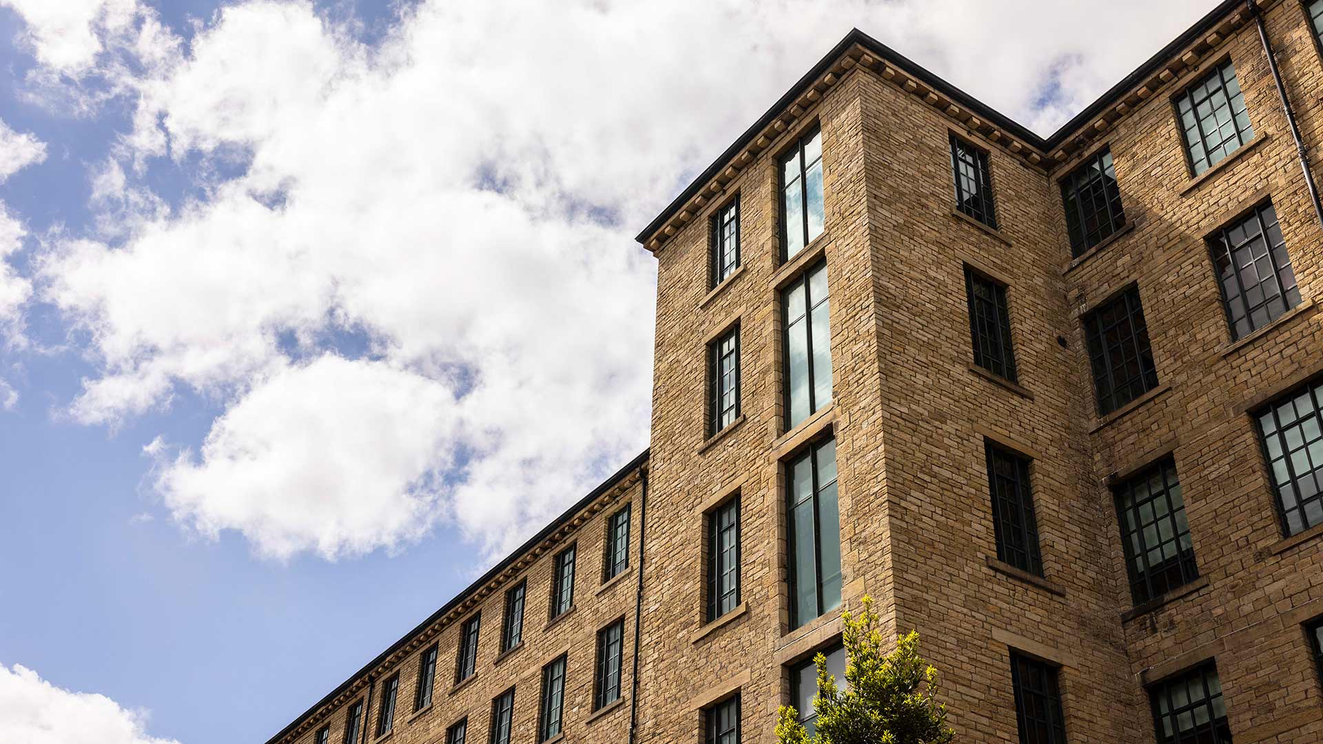 Converted mill university building with multiple storeys and large windows, viewed from below against a partly cloudy sky, with tree foliage visible in the foreground.