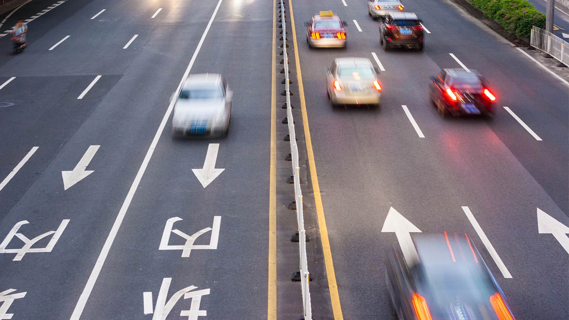Multi-lane road in China with moving traffic, showing cars blurred in motion, directional arrows, and road markings in Chinese characters separated by central barrier.