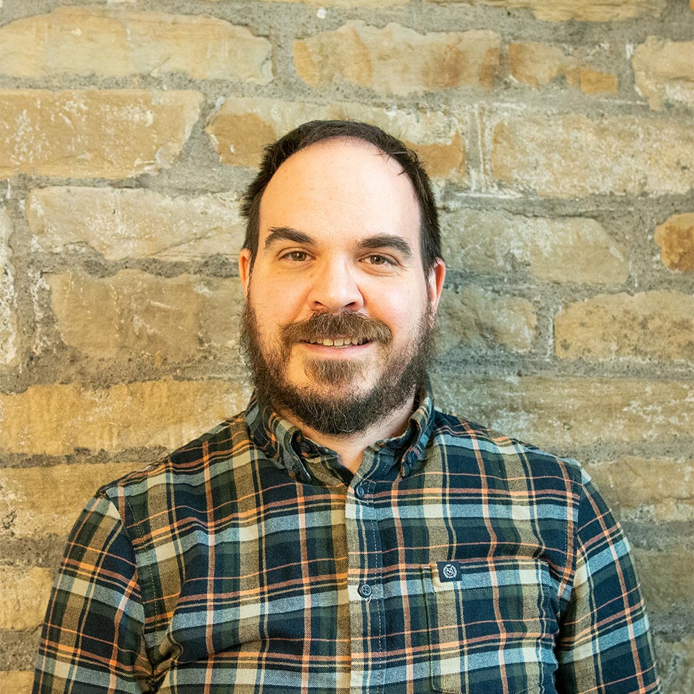 Head-and-shoulders portrait of an individual in a checked shirt standing in front of a stone wall.
