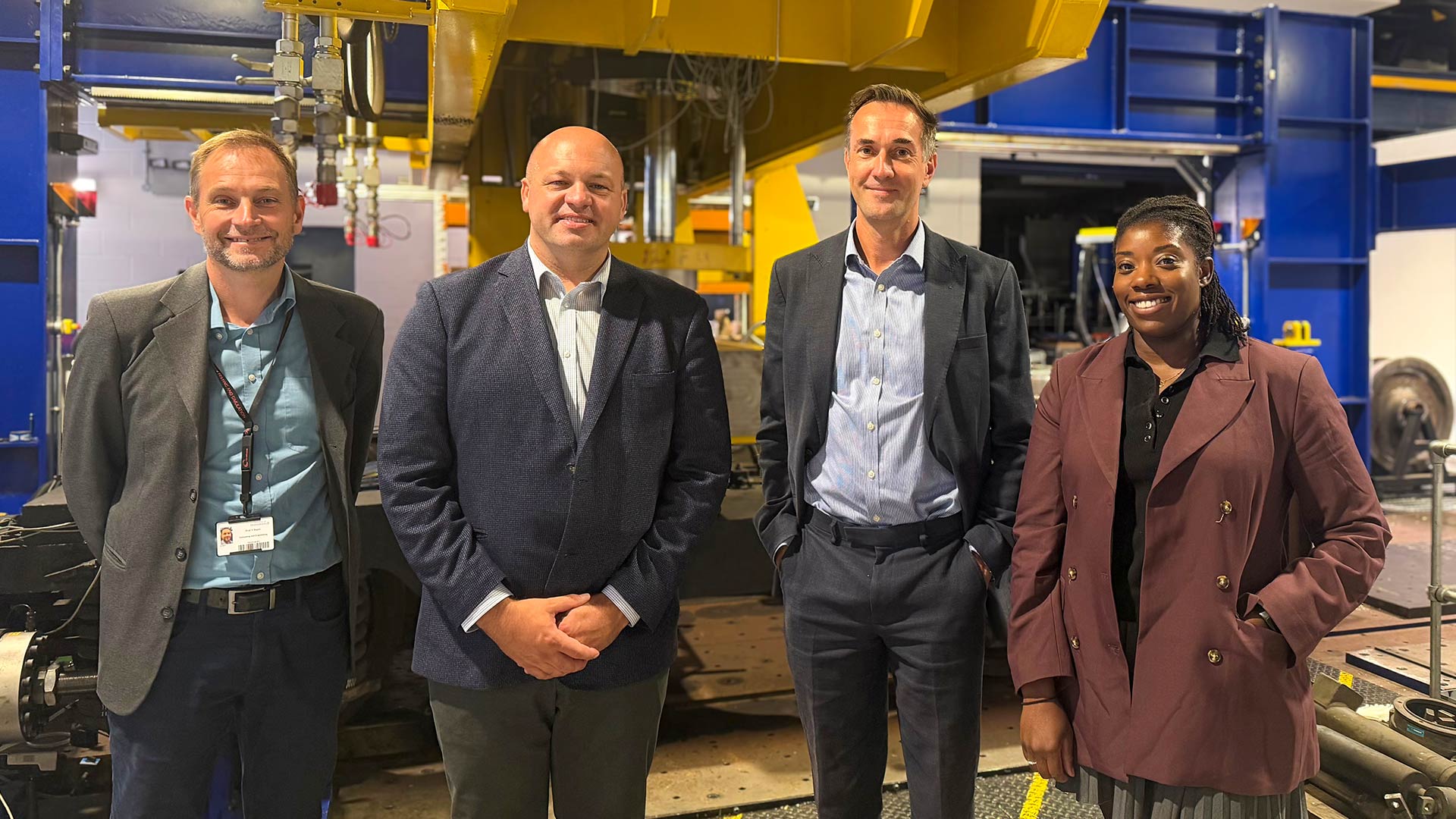 Four individuals stand beside large railway testing equipment in an engineering research facility, with blue and yellow industrial machinery and laboratory infrastructure visible in the background.