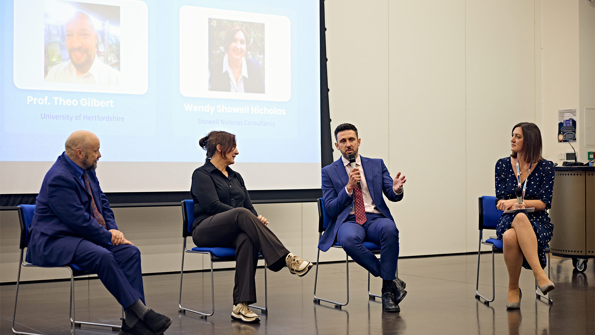 Panel discussion taking place in lecture theatre, with speakers seated on stage in front of presentation screen during health and social care event.