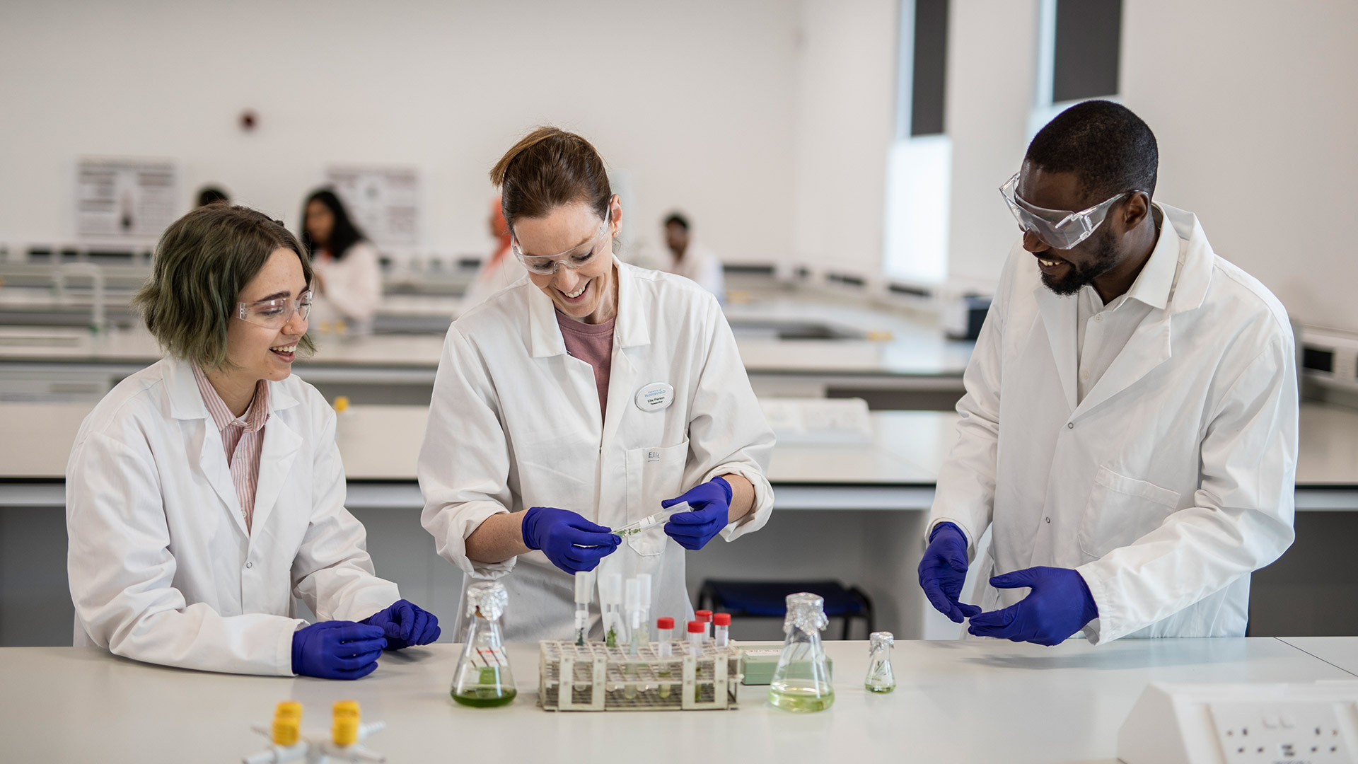 Three students in a lab, wearing lab coats, glasses and rubber gloves, working with chemicals and glass equipment.