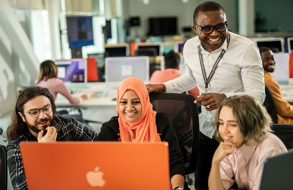 Three students working together, using a desktop computer device with an academic stood behind them.