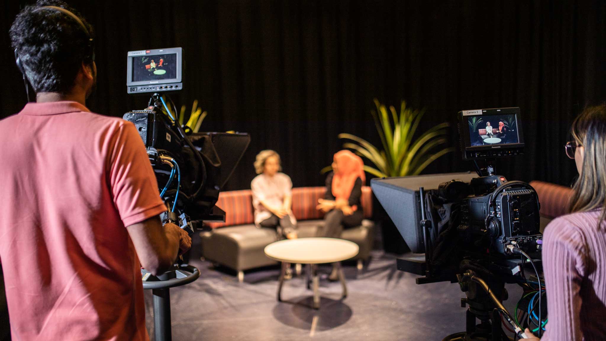 Students holding camera whilst other students chat on a sofa
