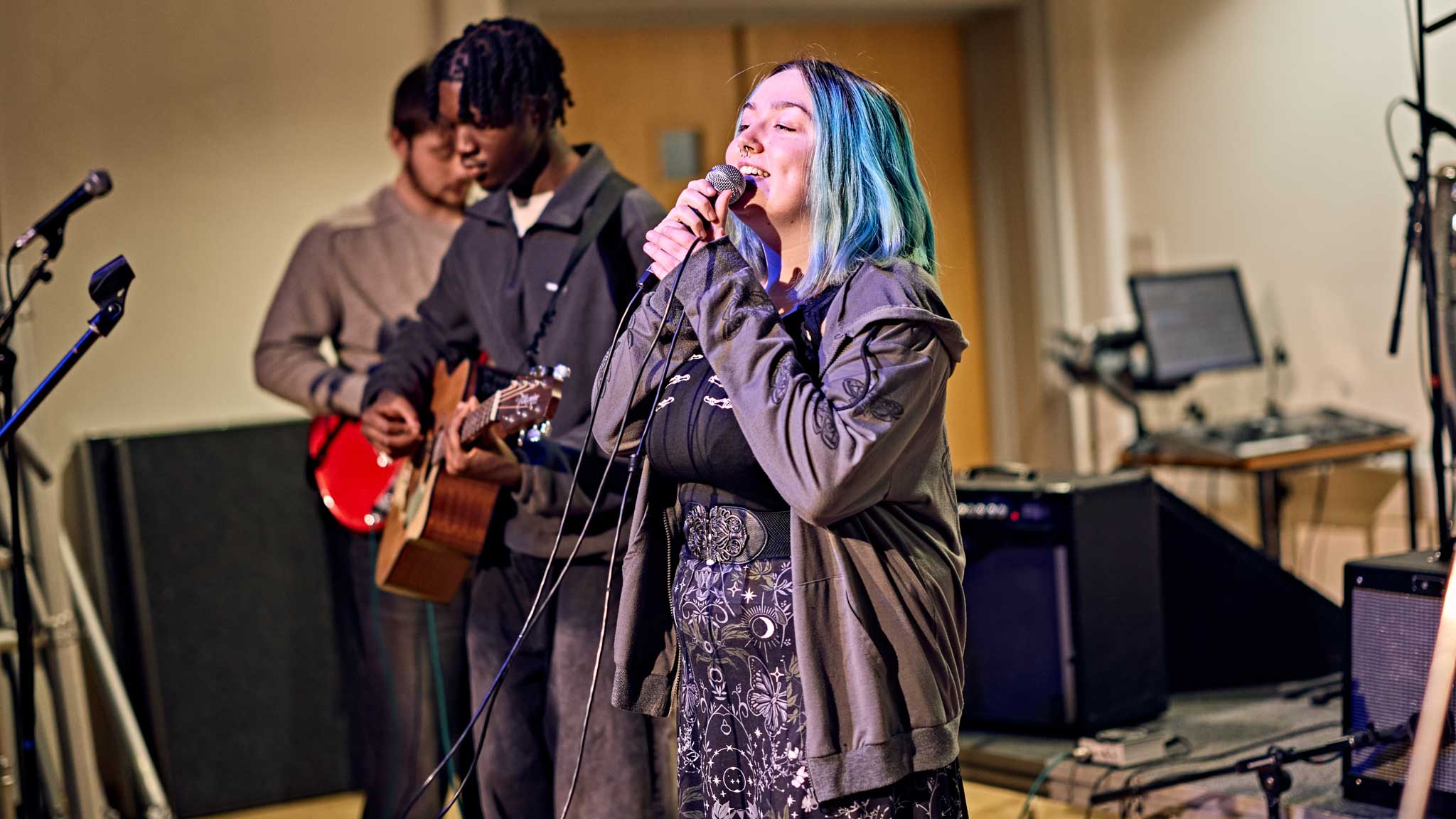 A student sings into a microphone whilst two other students play guitar