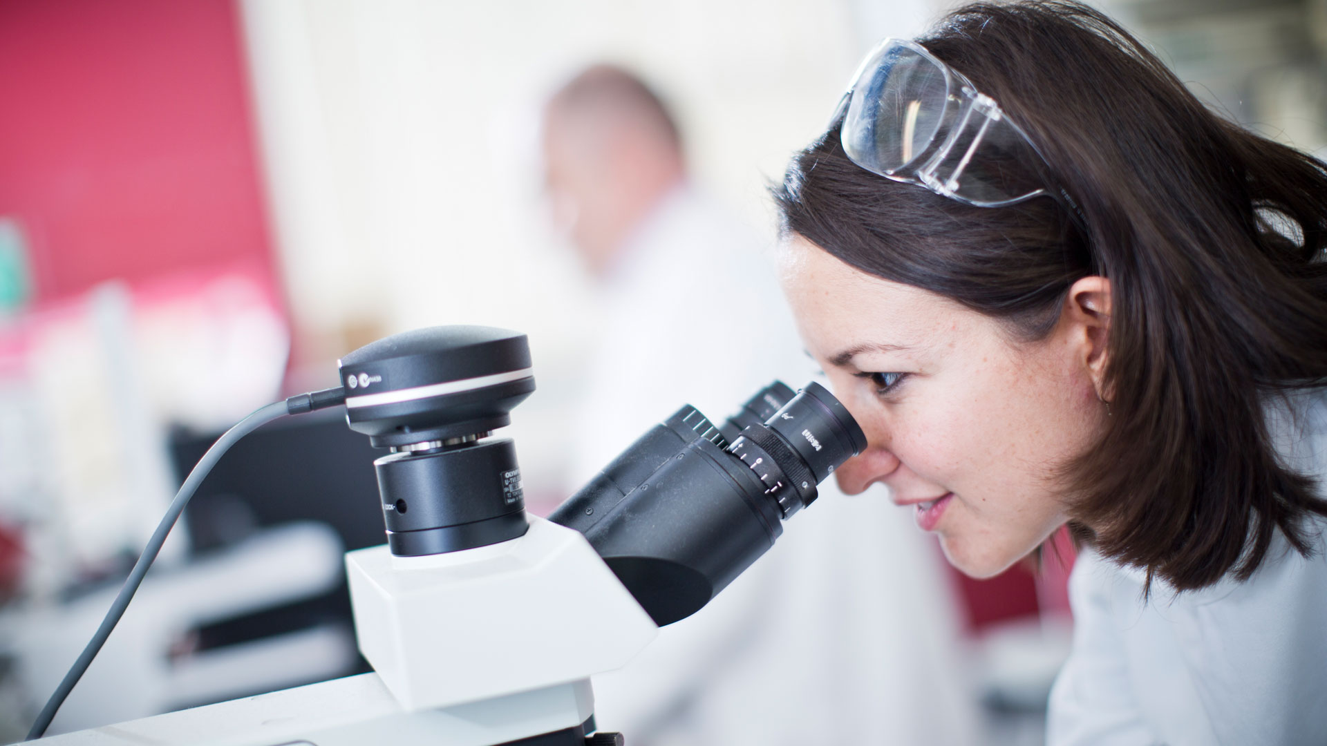 A researcher in focus peers intently into a microscope. Behind her another researcher is blurred out.