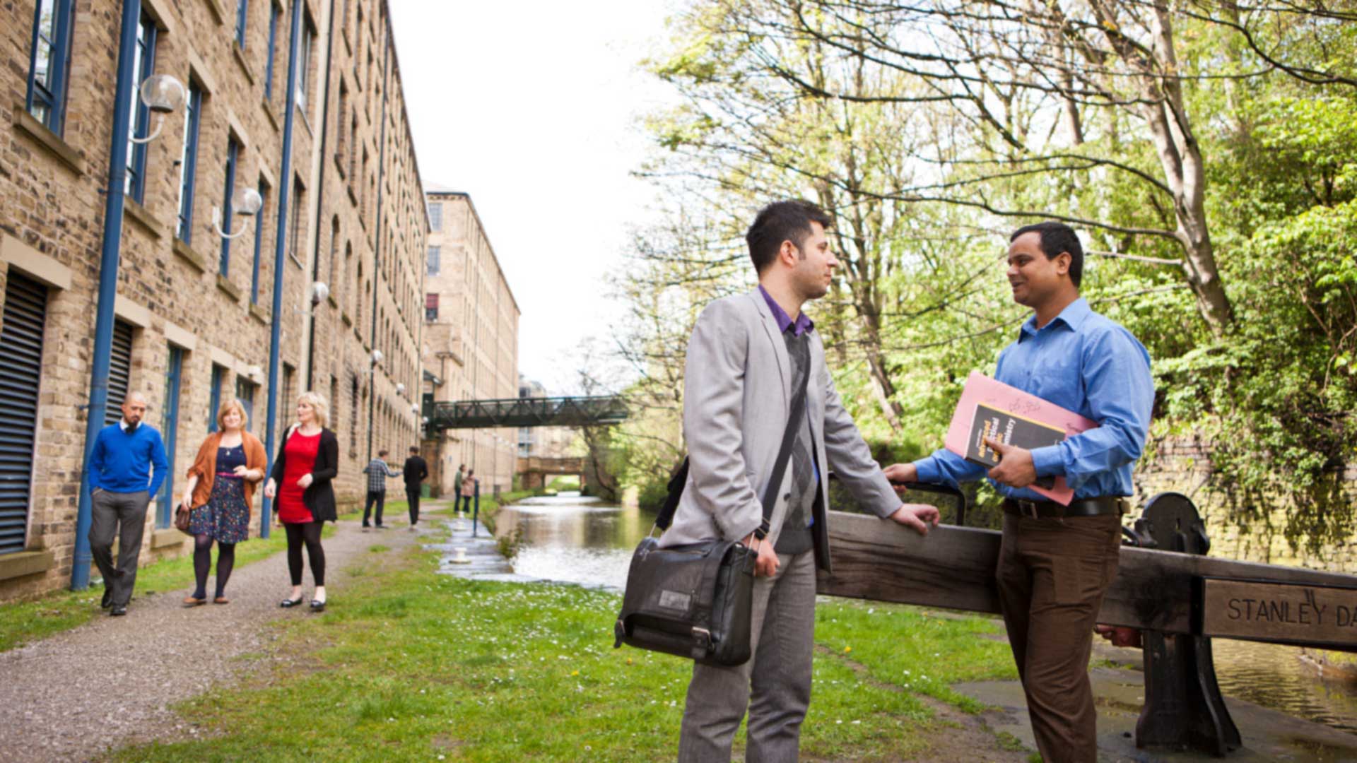 Two students stand chatting next to the canal behind one of the canalside buildings. Other students walk in the background.