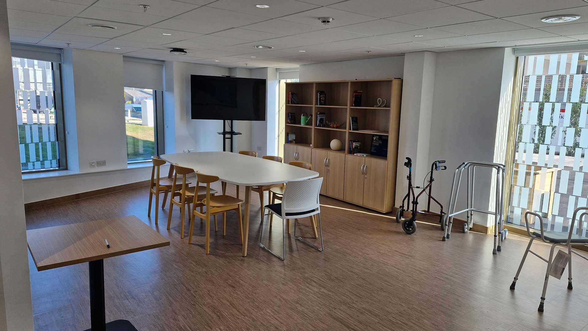 The dining room in the Community House facility in the Daphne Steele building on campus. There is a long table with chairs, shelving, a TV and various Zimmer frame/walking aids.