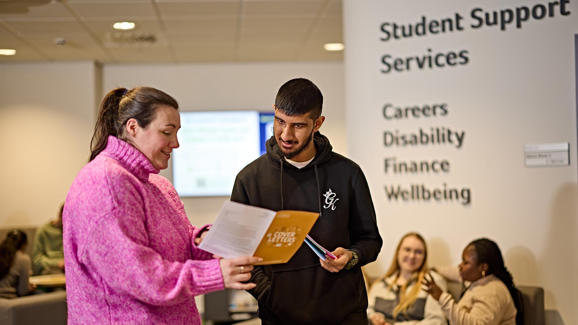 Edwin talking to a member of staff from one of the student support services, there is a wall behind them that lists the different services available.