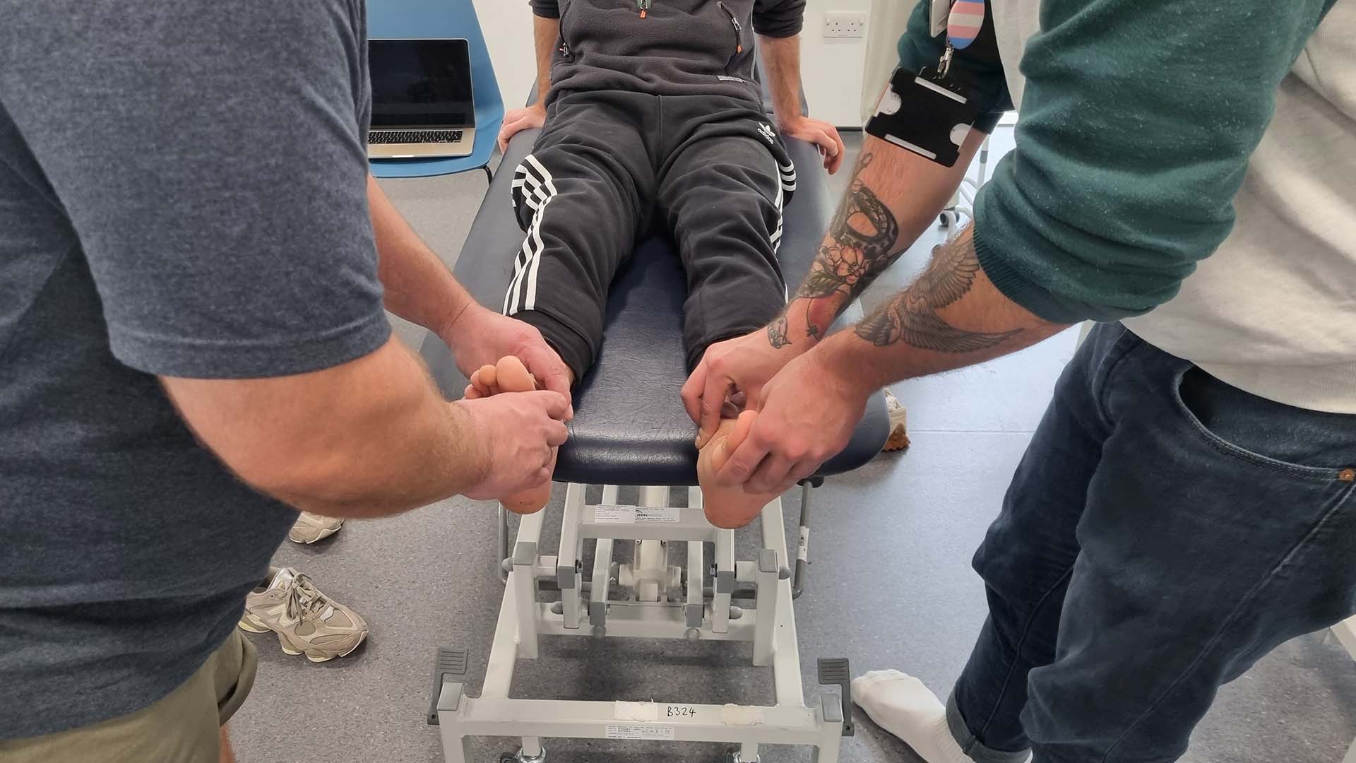 Two students practising podiatry on another student's bare feet, who is sitting on a bed.