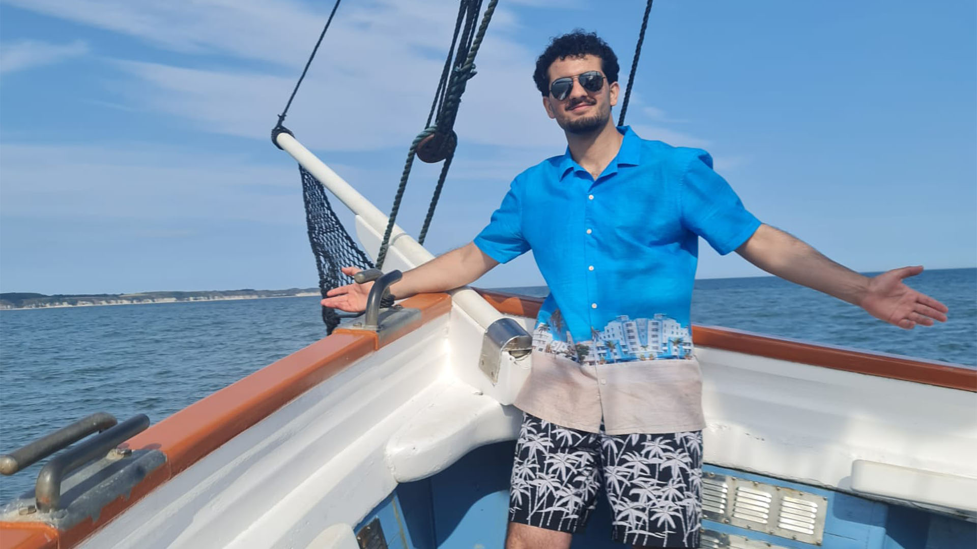 Saad Zahir standing on a boat, wearing bright, summery clothes, smiling.