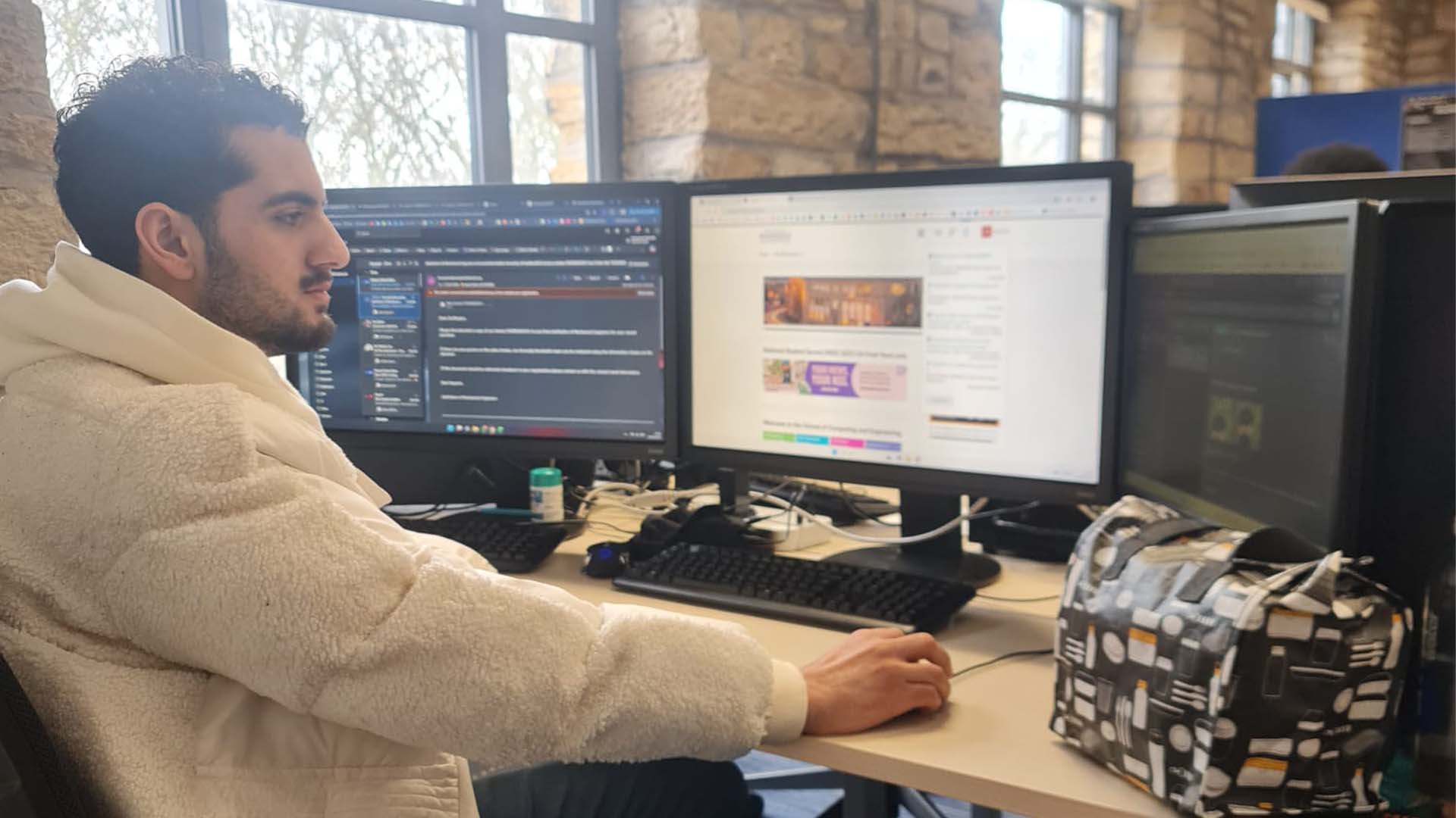 Saad Zahir sitting at a desk, using a computer with three screens, doing work.