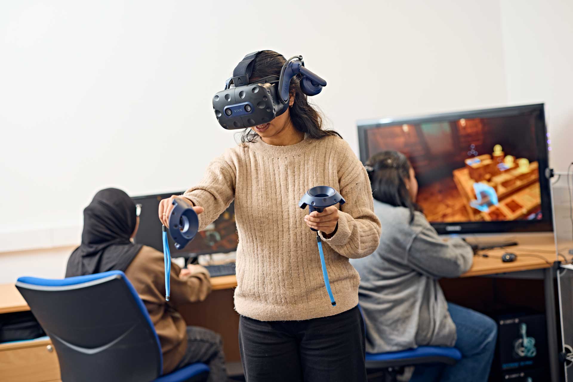 Three psychology students conducting an experiment using virtual reality equipment.  One student navigates a virtual reality environment using a VR headset while the other two monitor their behaviour on computer monitors