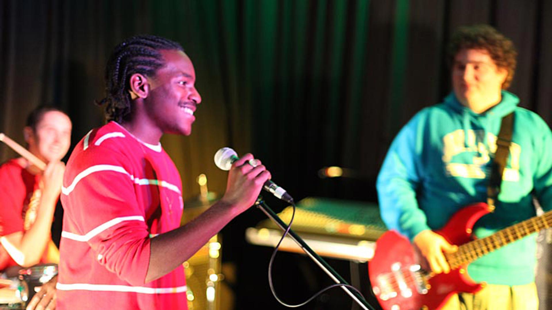 Image of someone singing into a mic while two other play instruments in the background