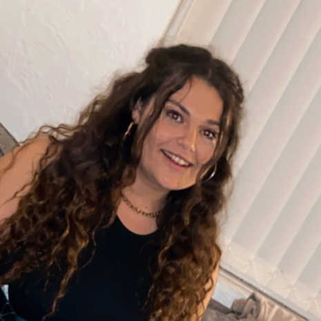 Hannah has long brown wavy hair and is wearing silver earrings and a black top