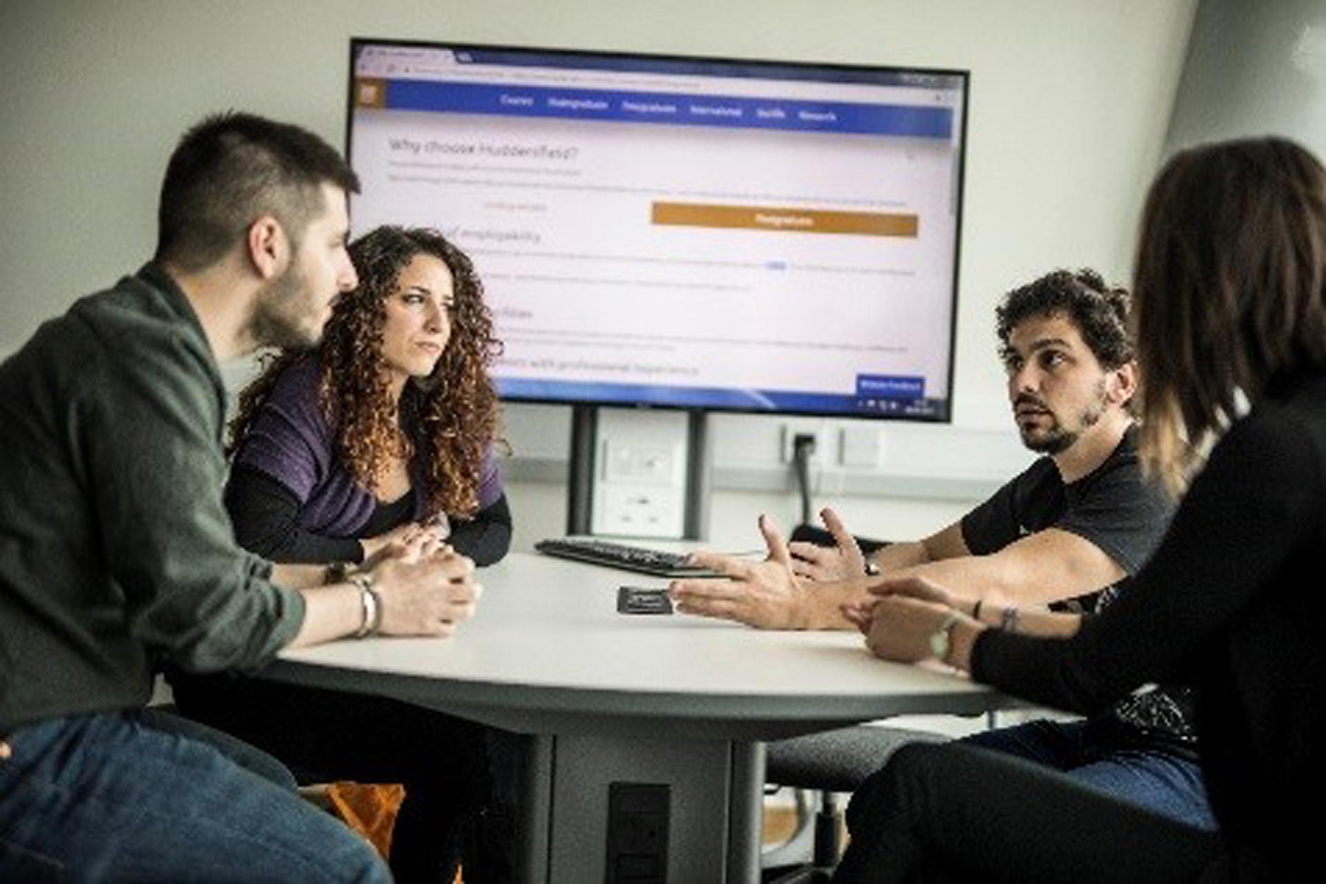 Three people talking around a table with the University of Huddersfield course finder site on a television in the background